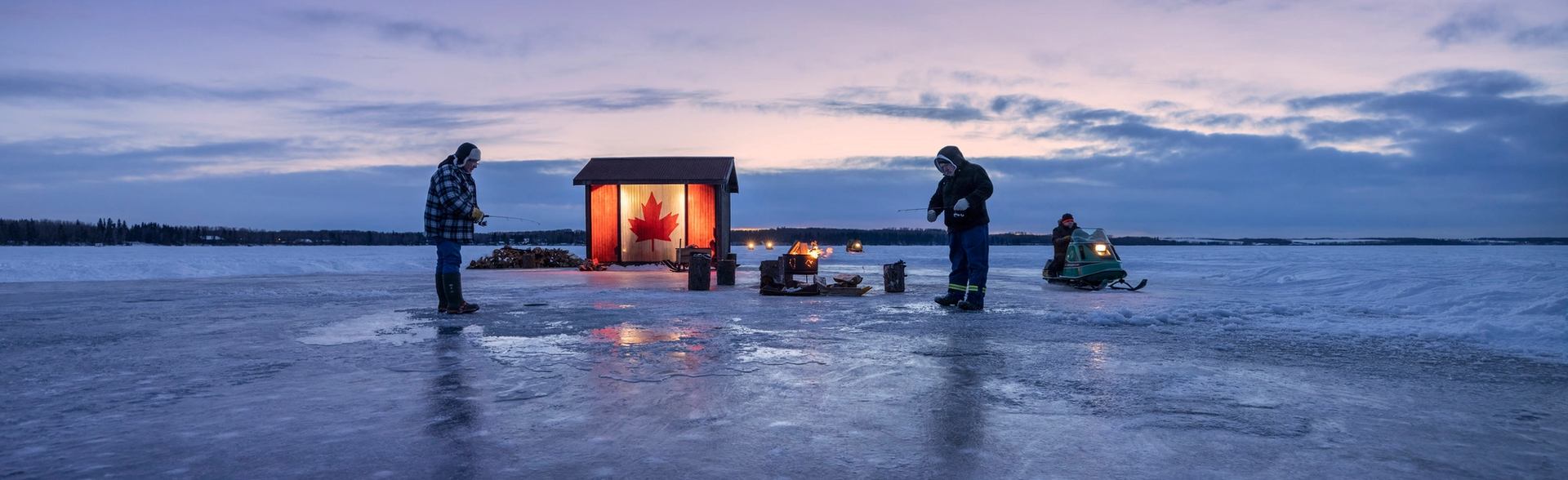 Two people ice fishing at sunset with an ice hut nearby.