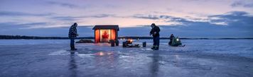Two people ice fishing at sunset with an ice hut nearby.