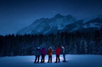 Four friends and a guide in snowshoes looking up at the night sky featuring the forest, mountains and stars