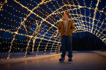 A skater pauses under a festively lit arch over an ice skating trail at night.