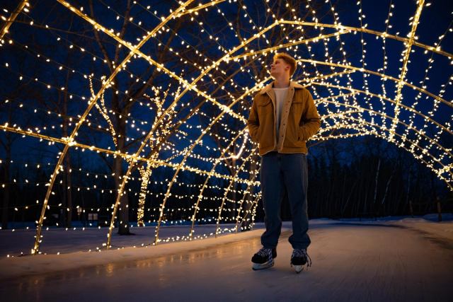 A skater pauses under a festively lit arch over an ice skating trail at night.