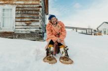 A Métis knowledge keeper sits outside a historical cabin in the snow, smiling widely and wearing traditional snowshoes.