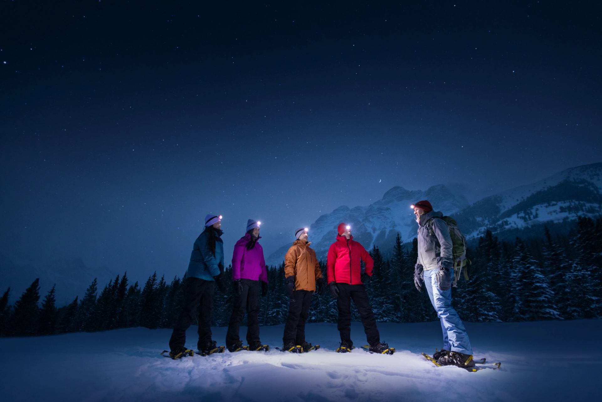 Four friends and a guide in snowshoes looking up at the night sky featuring the forest, mountains and stars.