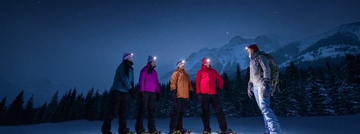 Four friends and a guide in snowshoes looking up at the night sky featuring the forest, mountains and stars.