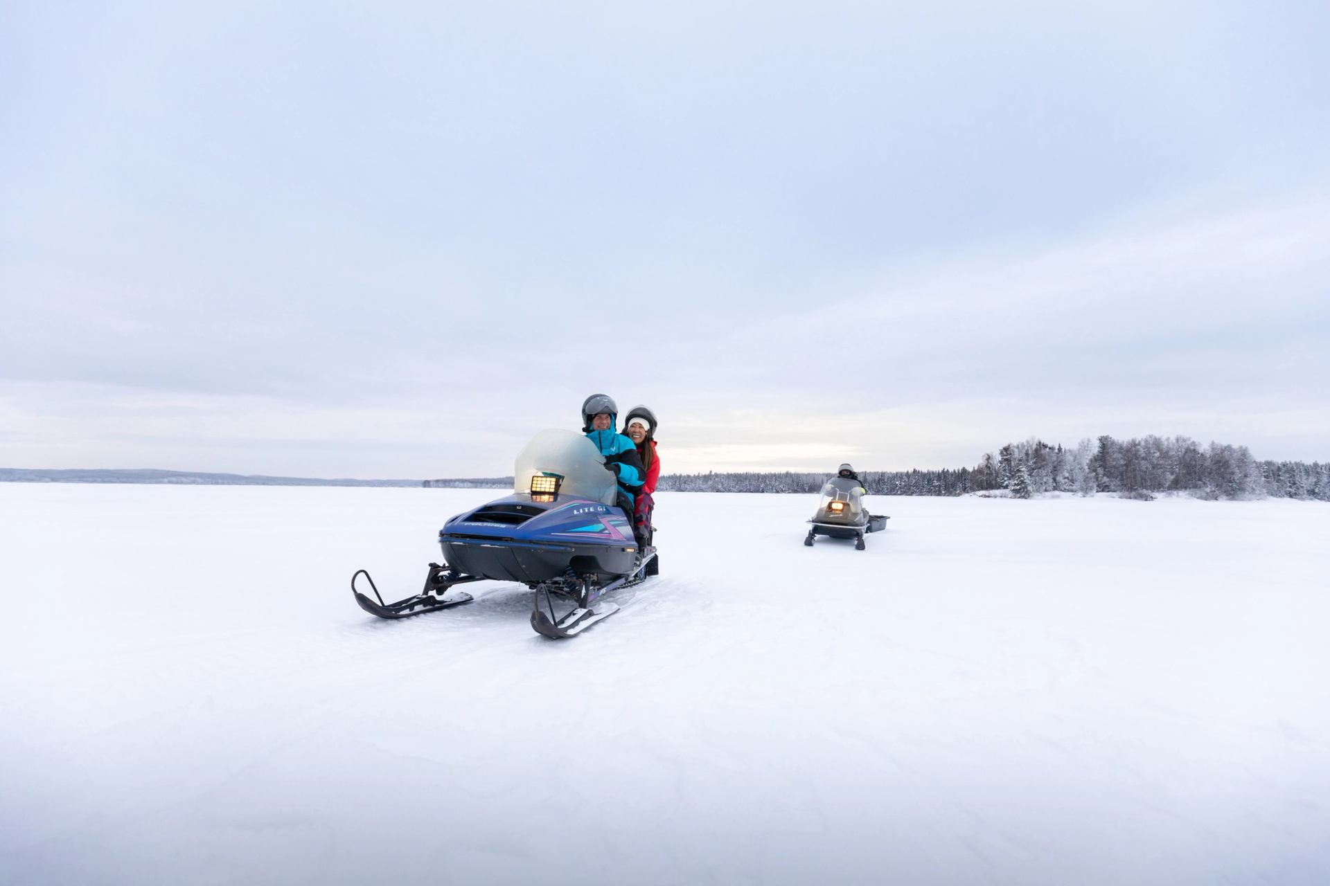 Couple snowmobiling on a lake, guide behind them, at Aurora Borealis Indigenous Village in Fort McMurray.