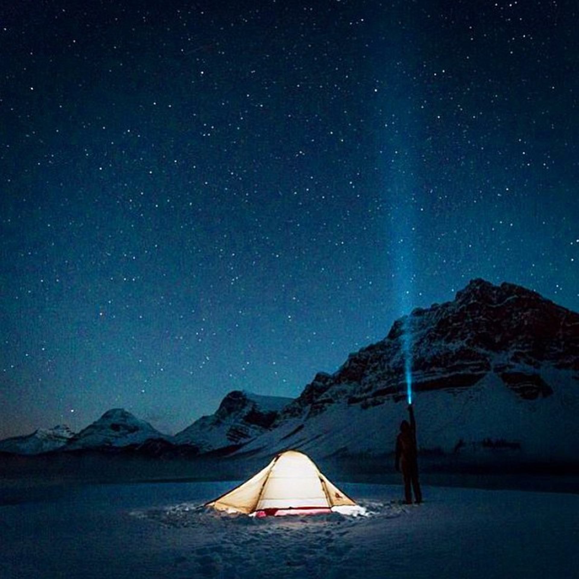 A person beside a tent at night, shining a headlamp into the dark starry sky while winter camping in the mountains.