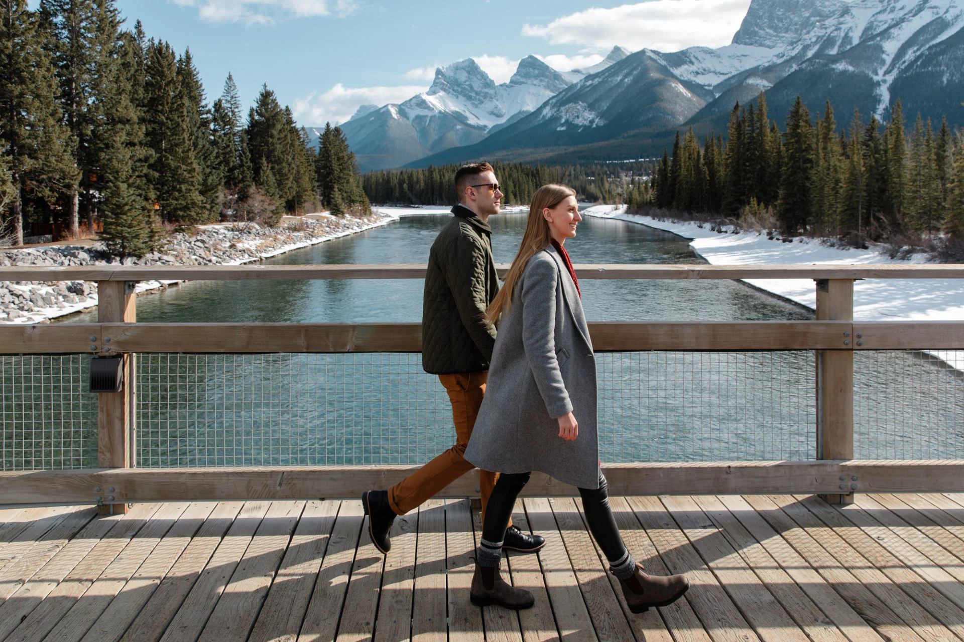 Couple walking along a riverside bridge with mountains in the background