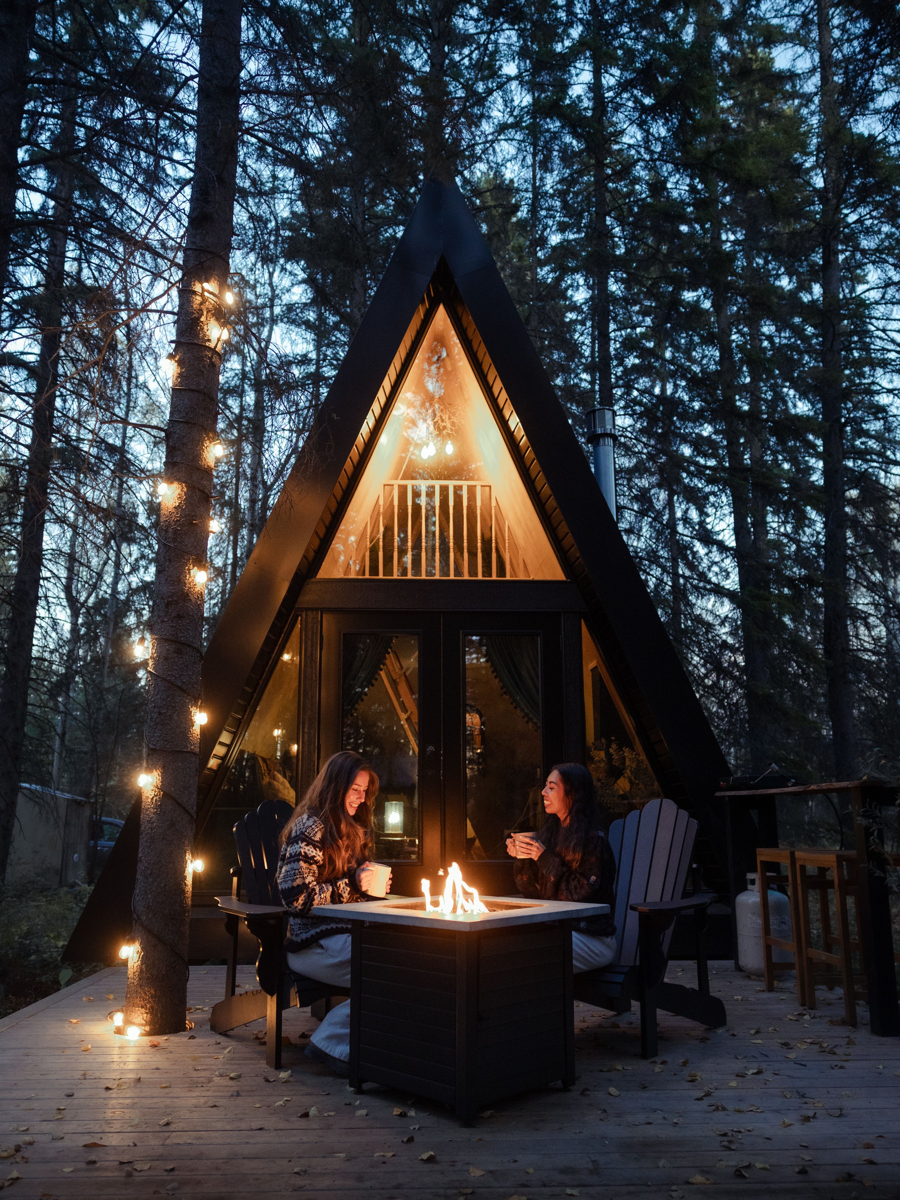 Two travellers sip warm drinks around a fire outside of a small A-frame cabin in the forest.