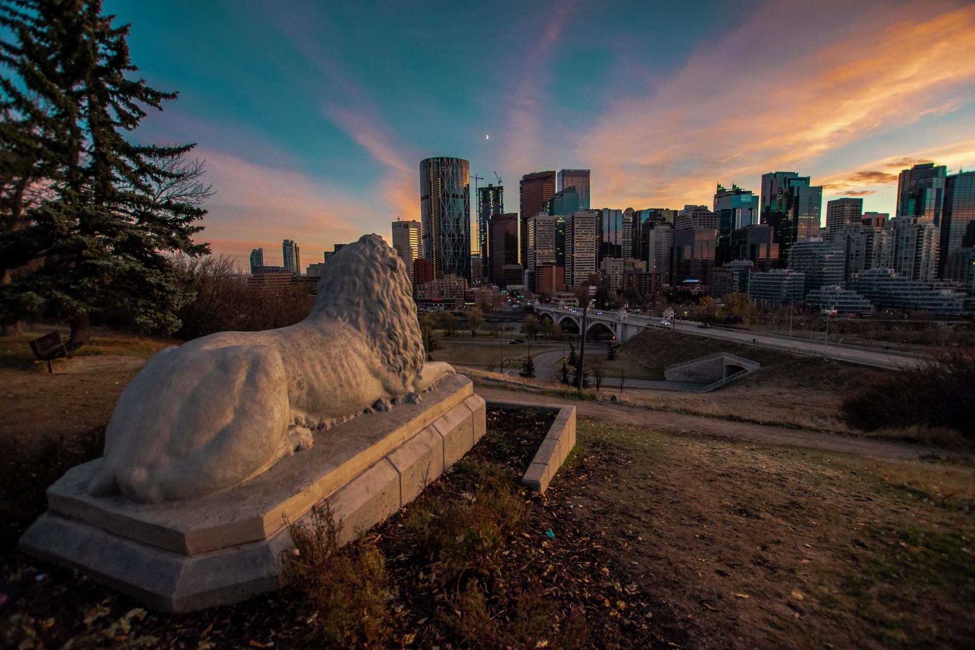 A large lion sculpture looks toward the Centre Street Bridge and downtown Calgary at sunset.