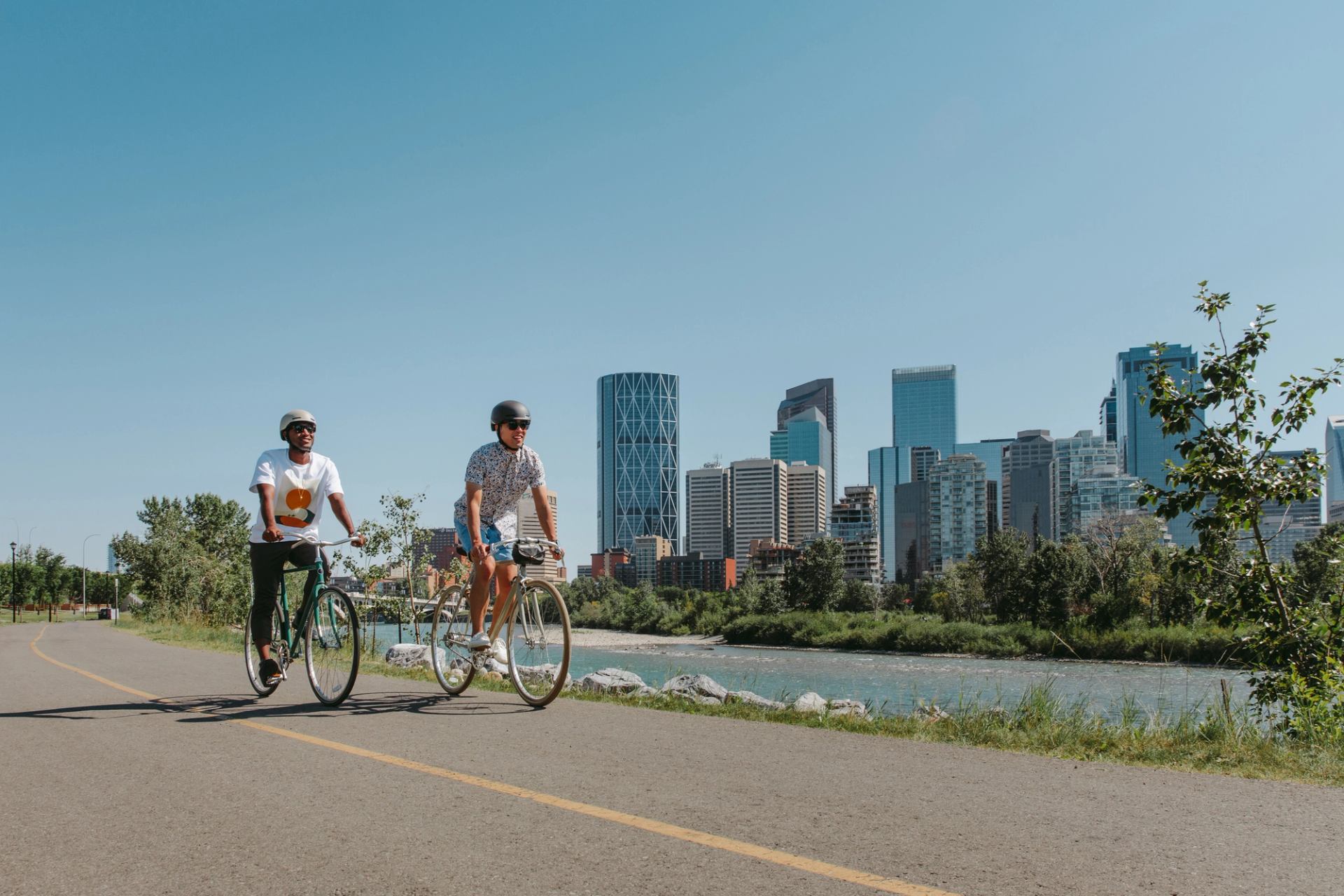 Two cyclists ride bikes on a paved riverside path with downtown Calgary nearby.