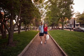 Couple walks along a tree-lined path with a shopping bag and a coffee.