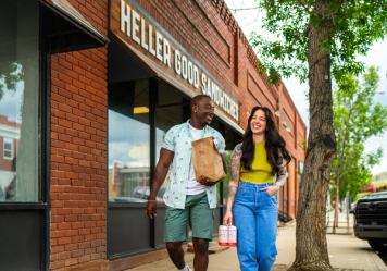 Two people take sandwiches to go as they walk away from the Heller Good Sandwiches storefront.