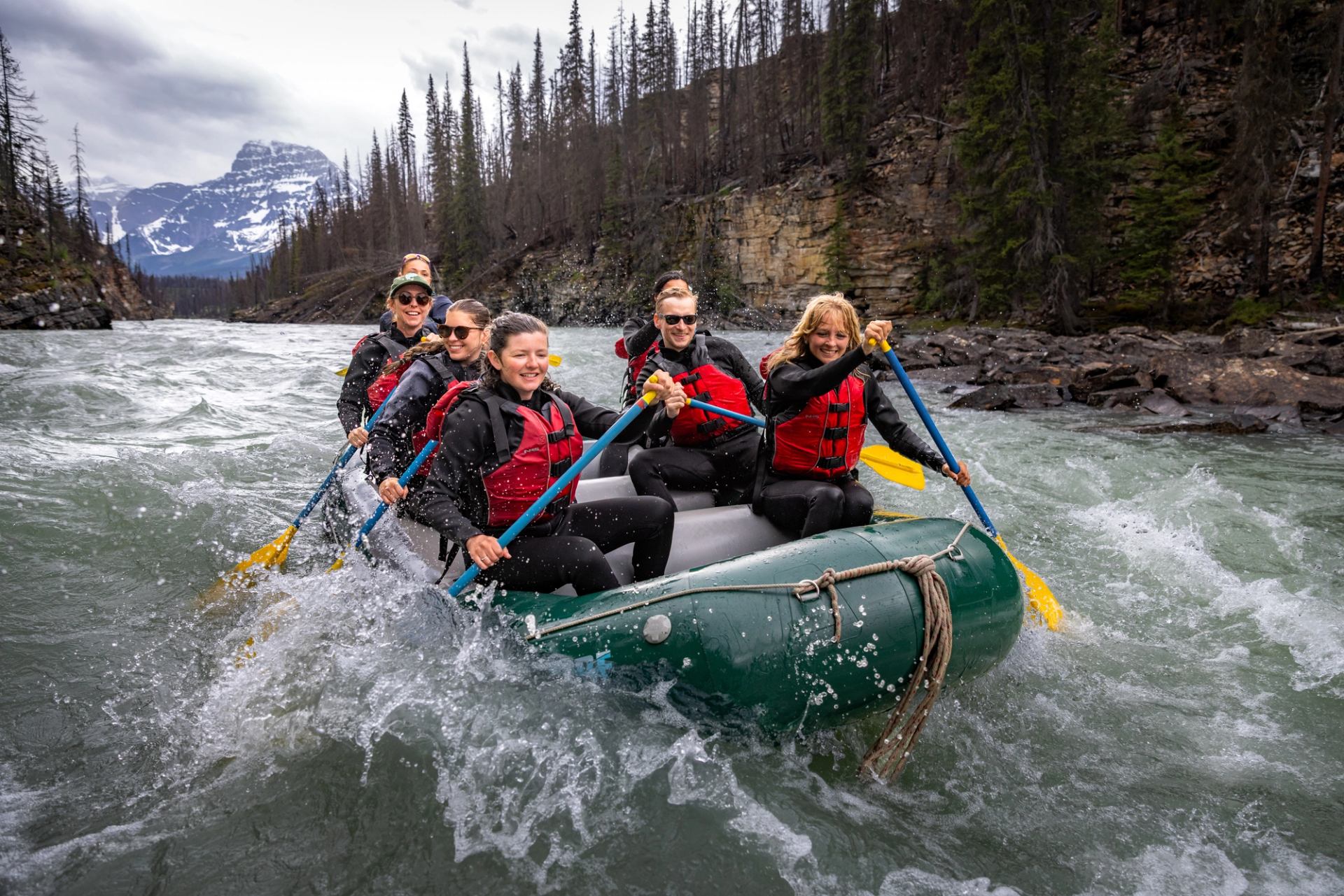 A group paddles through rapids on a river with mountains in the distance and trees along a mountainous shore.