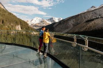 Two male kids standing along a clear glass railing of a skywalk overlooking a mountain view.