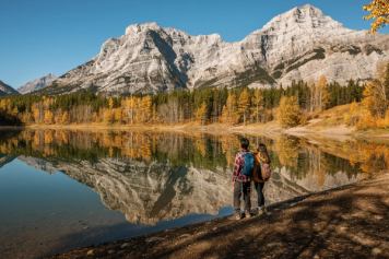 Two people stop at an alpine pond with Rocky Mountains in the nearby distance.