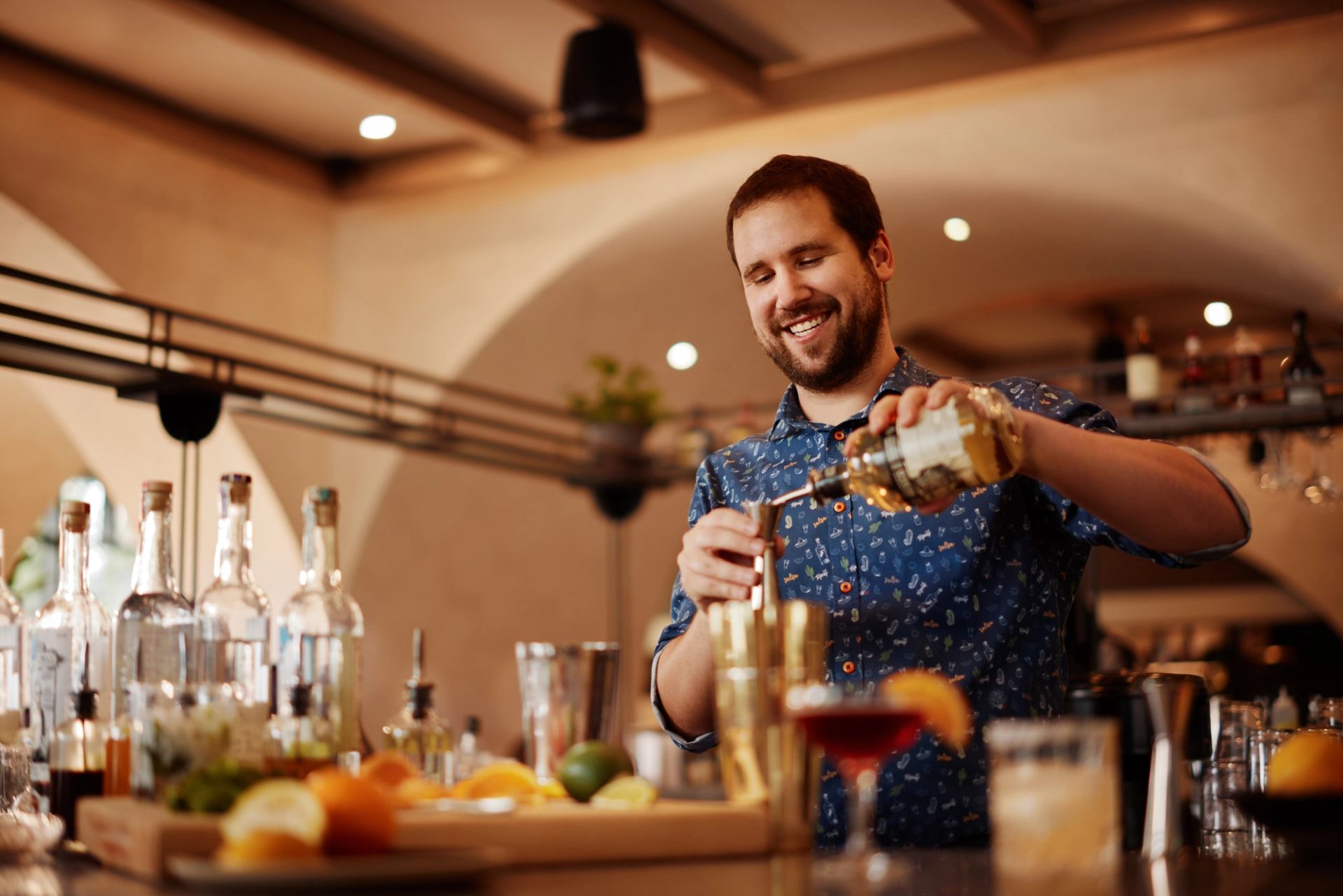 Smiling barman pours a measure of alcohol in an amply stocked bar while an inviting cocktail sits on the bar counter