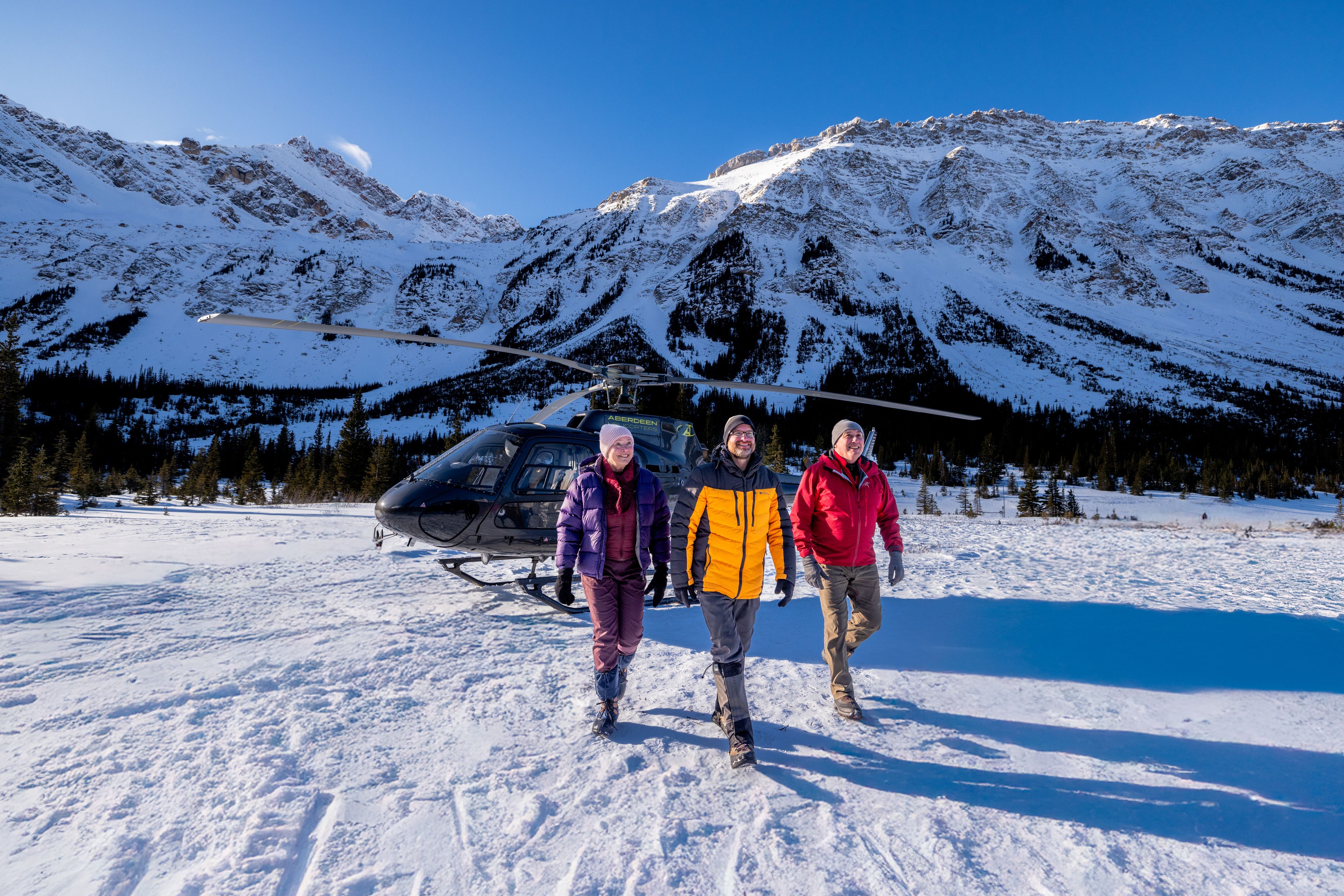 Three people walk away from a helicopter landed among snowy mountains.