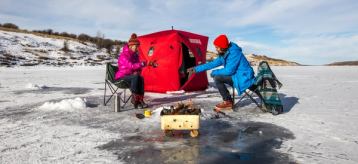 A couple sitting in front of a portable ice hut, with a fire going while ice fishing.