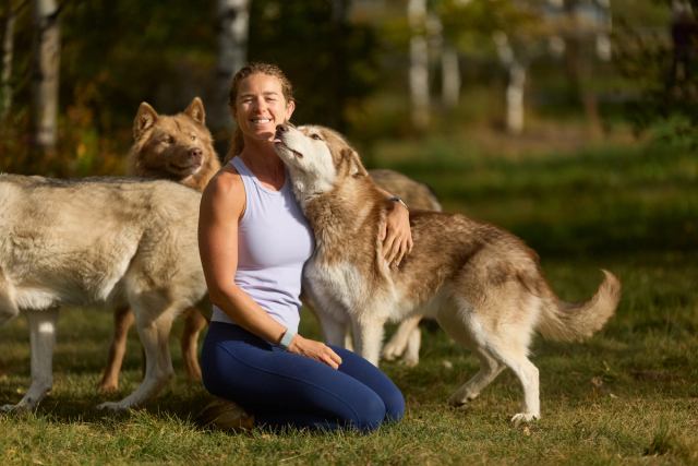 The owner of Yamnuska Wolfdog Sanctuary posted with a pack of young wolfdogs while one licks her face.