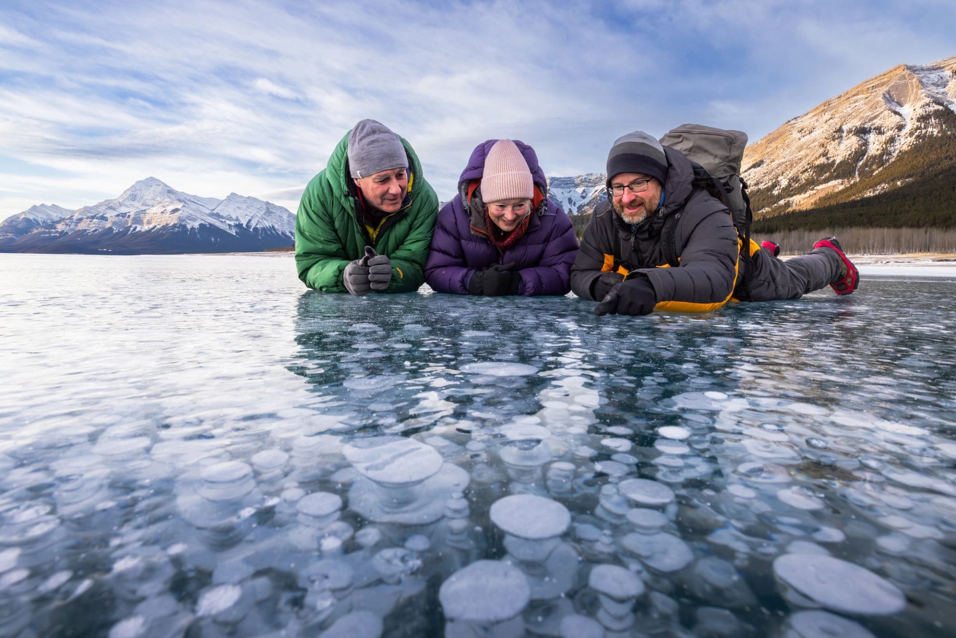 A couple with a guide laying on ice observing ice bubbles at Abraham Lake.