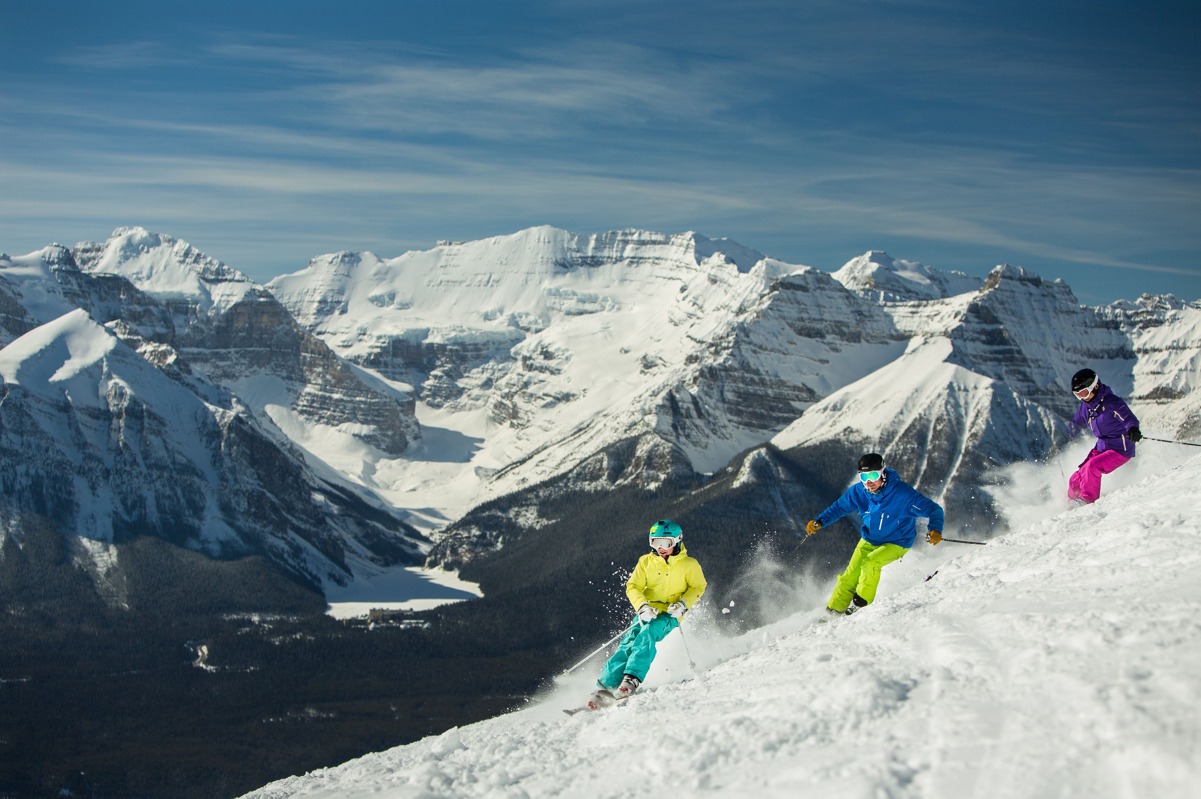 Three skiers ski down a mountain with snowy summits in the background.