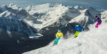 Three skiers ski down a mountain with snowy summits in the background.
