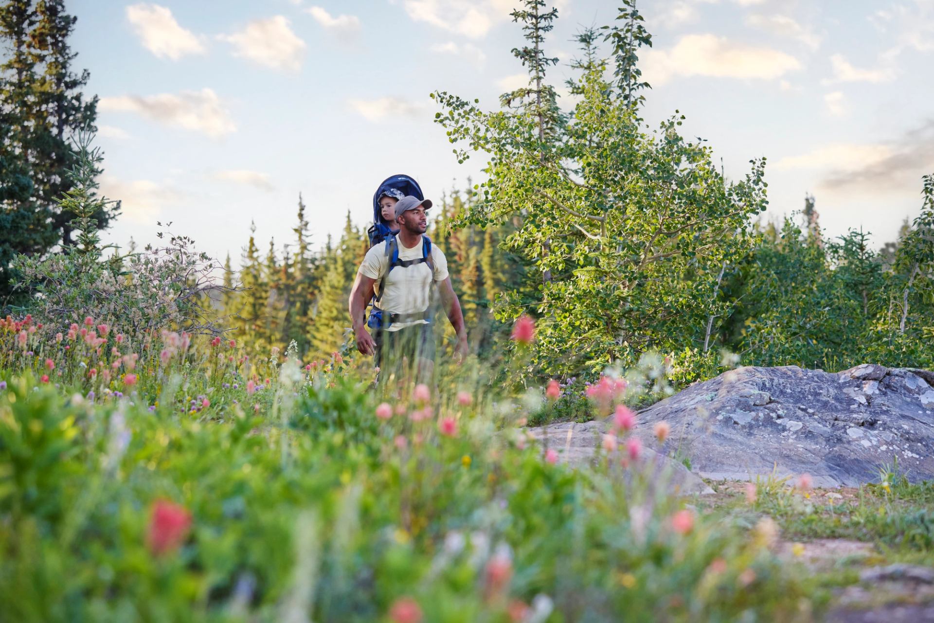 A man with a child on a hike with pink flowers and lush greenery near the path at the Athabasca Lookout. 