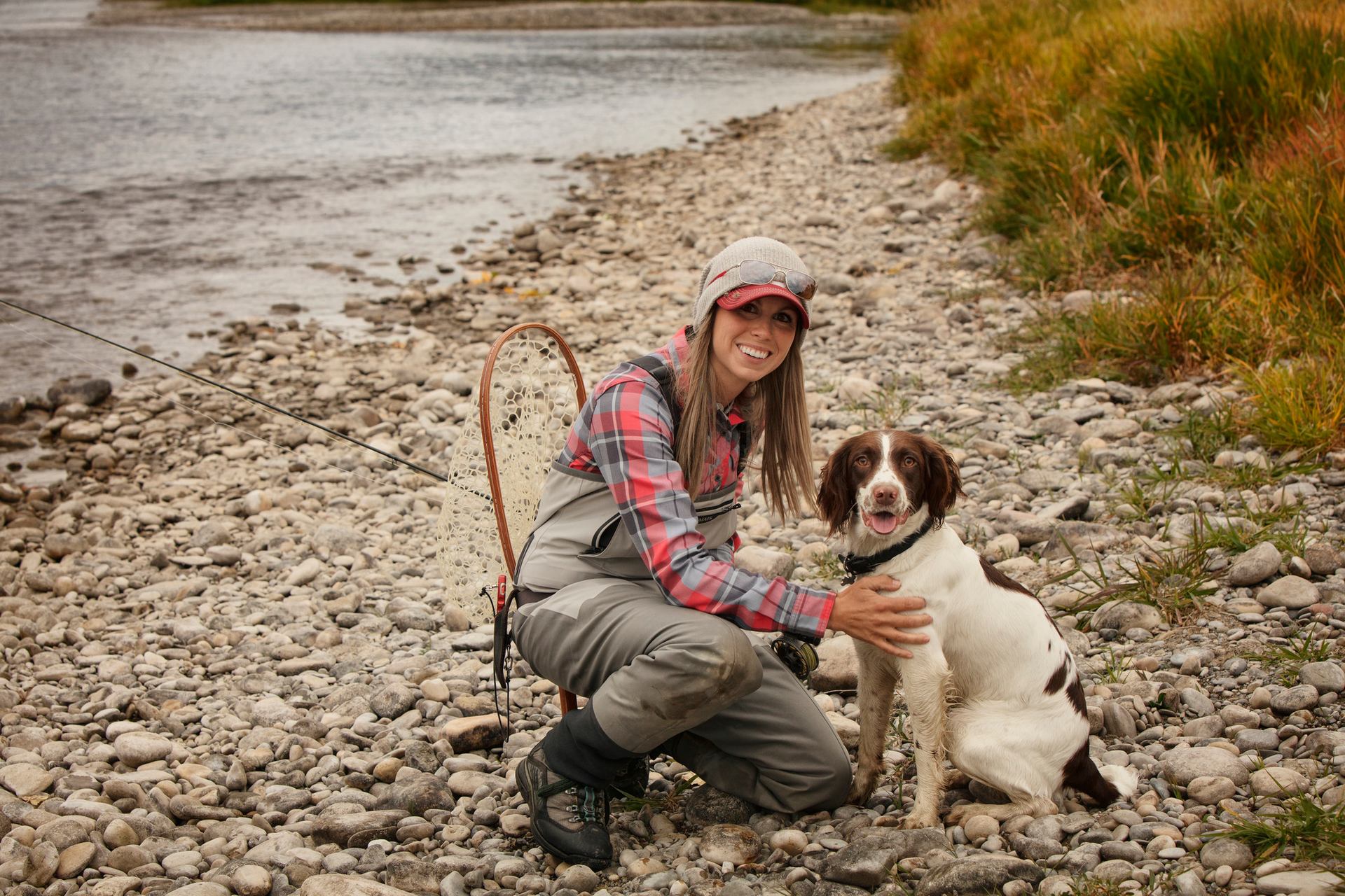 A woman and her dog pose by the river while out fly fishing.