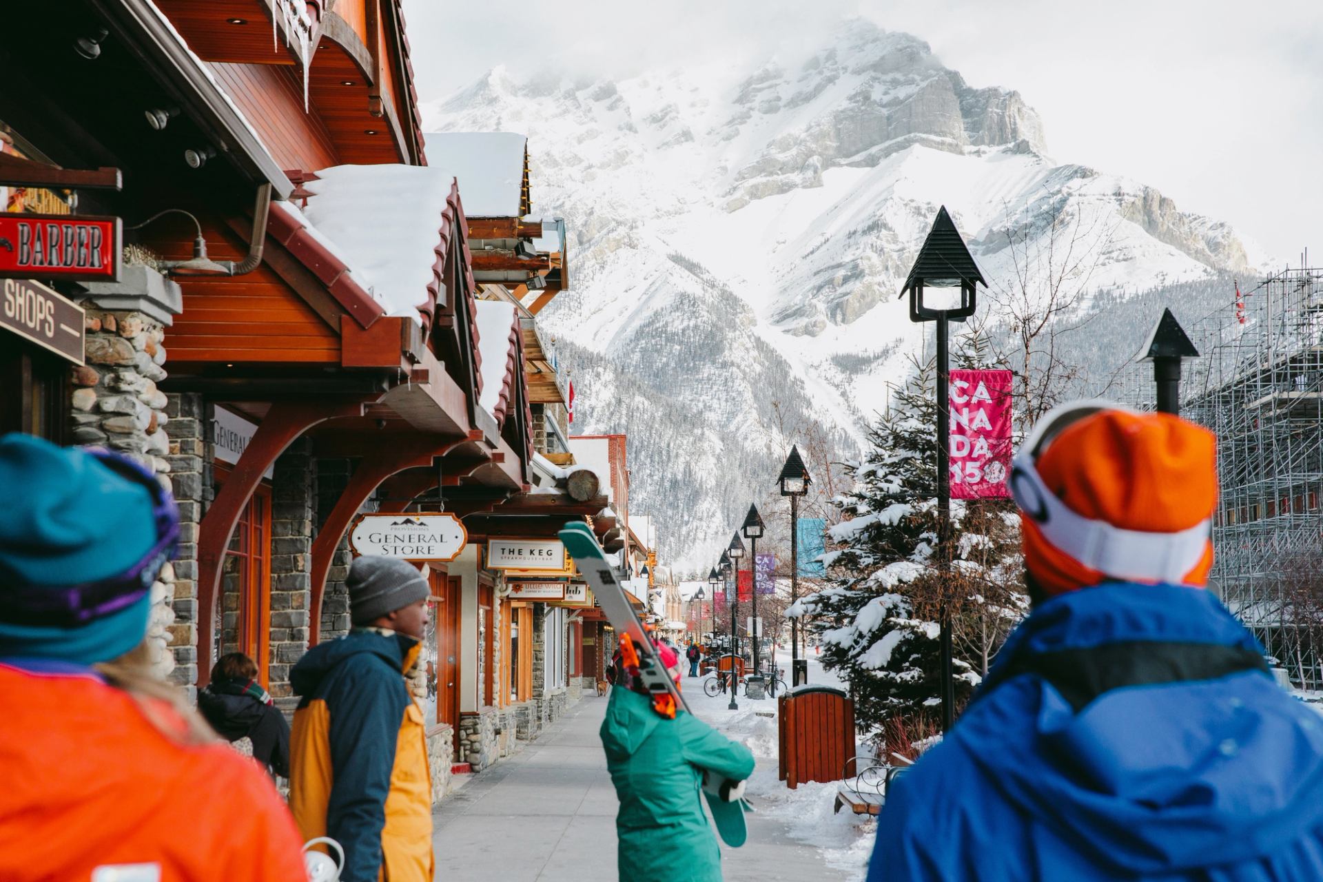Up close from behind, a group of skiers and snowboarders walking down the street, with the town and mountain in the background, located within Banff National Park