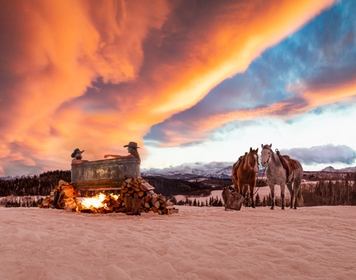 Couple in outdoor rural hot tub in winter during a sunset with horses in the background.