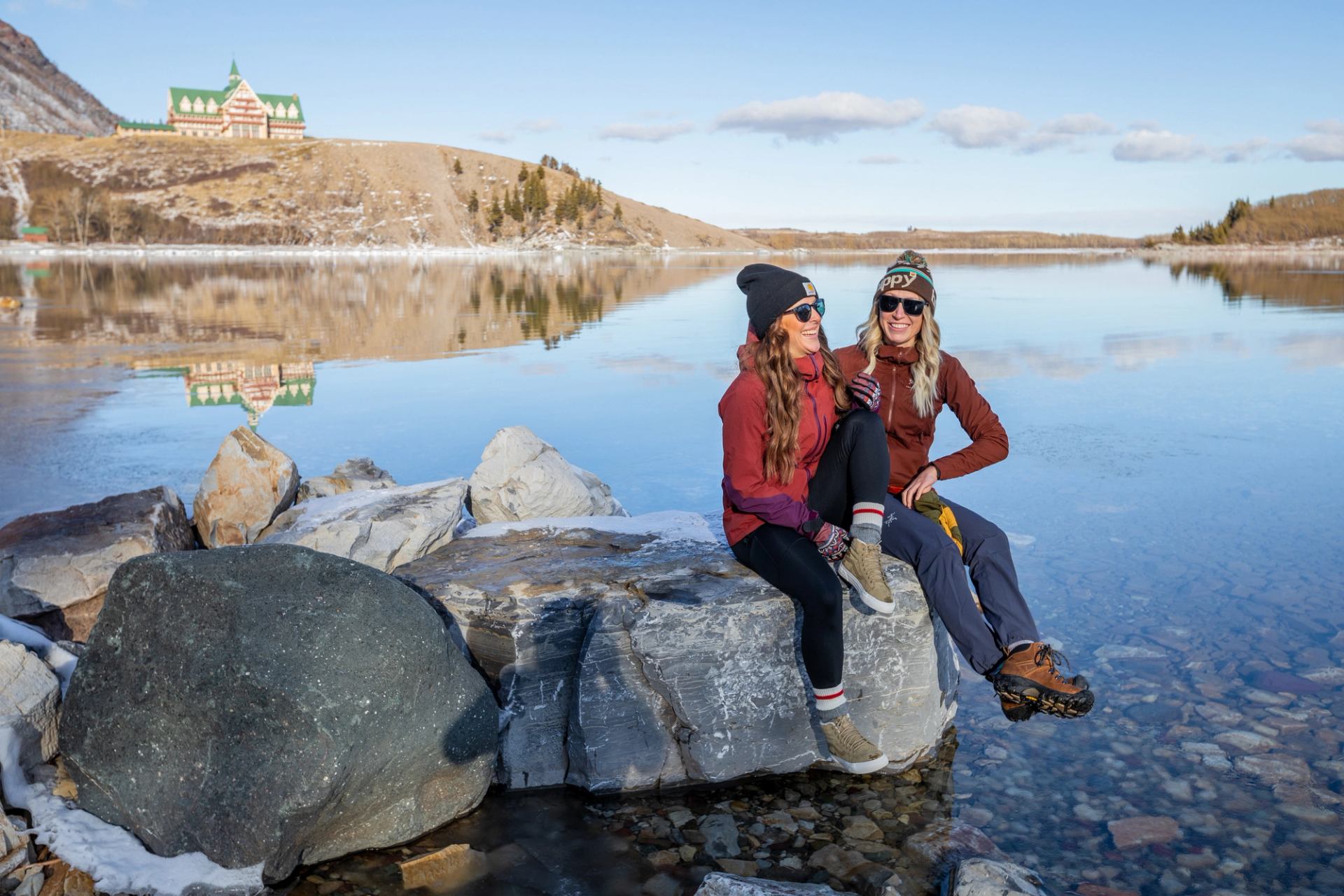Two women sit and talk on a rock in the water with the Prince of Wales Hotel in the background, in Waterton Lakes National Park