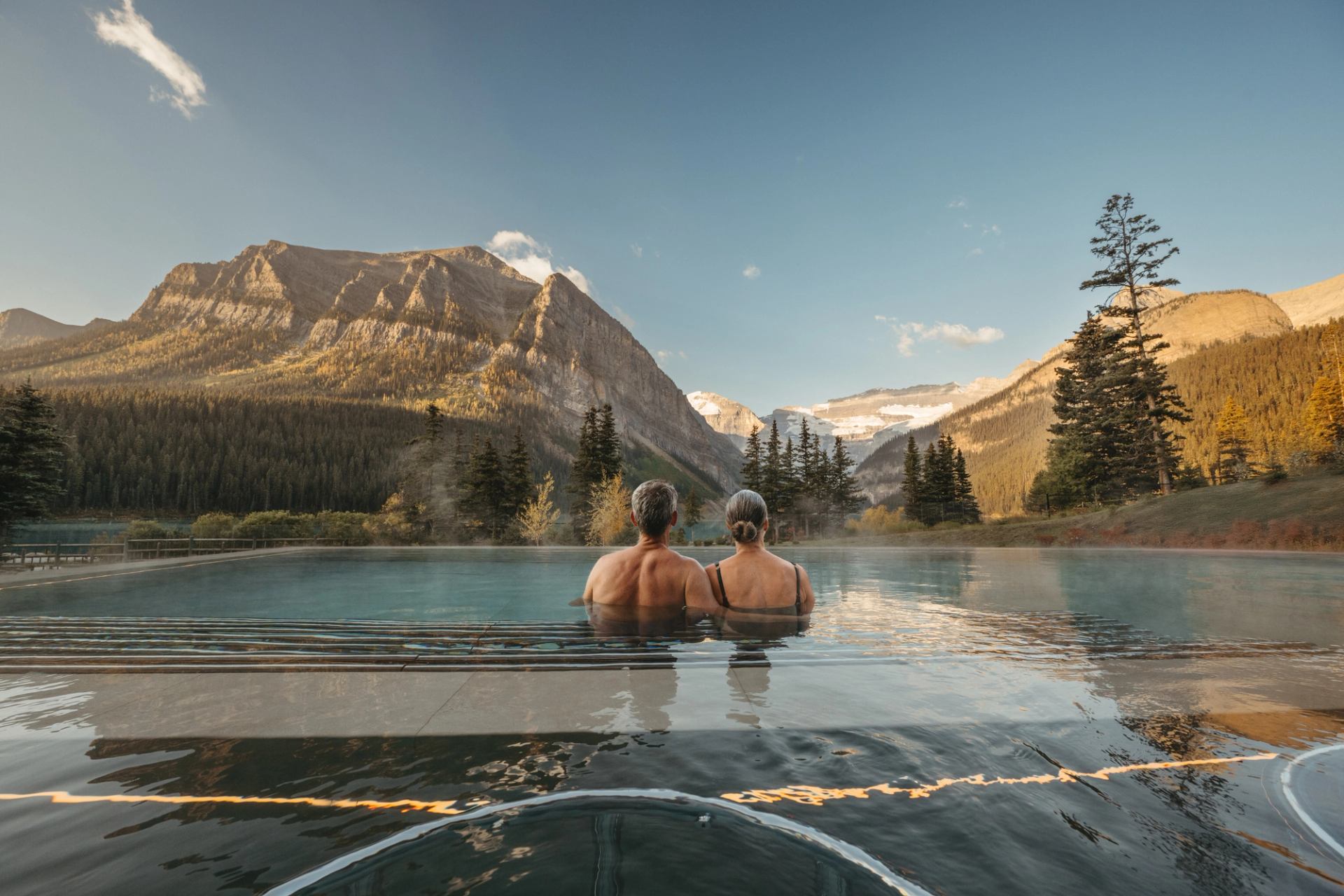 Two people soak in an infinity pool looking toward the nearby Canadian Rockies at BASIN Glacial Waters in Lake Louise.