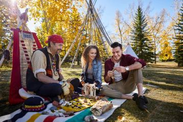 Couple sitting with an Indigenous craftsman near teepee poles at Heritage Park, admiring his works on a Hudsons Bay blanket