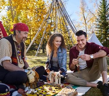 Couple sitting with an Indigenous craftsman near teepee poles at Heritage Park, admiring his works on a Hudsons Bay blanket