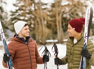 Two people with cross country skis on a snowy trail near a stand of trees.