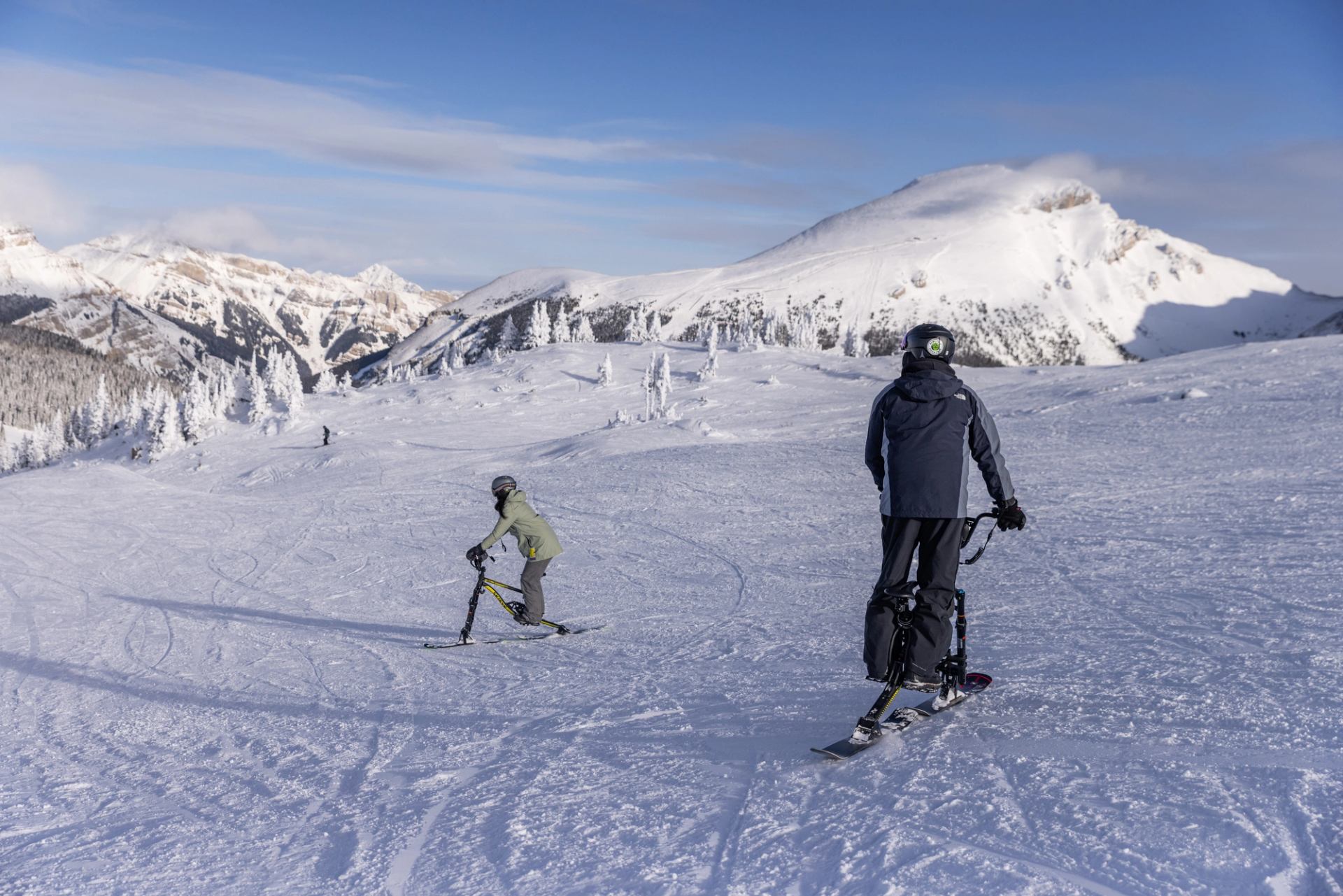 Two people ski biking on bikes with skis instead of wheels, surrounded by mountains at Banff Sunshine Village.