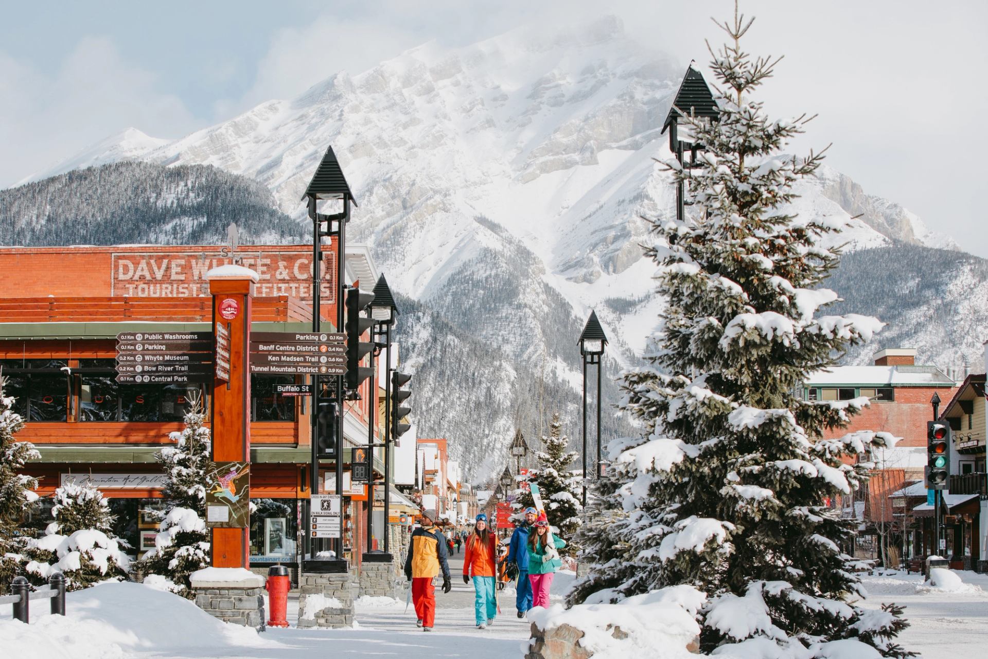 From behind, a group of skiers and snowboarders walking down the street with the town and mountain emphasized in the background, located within Banff National Park.