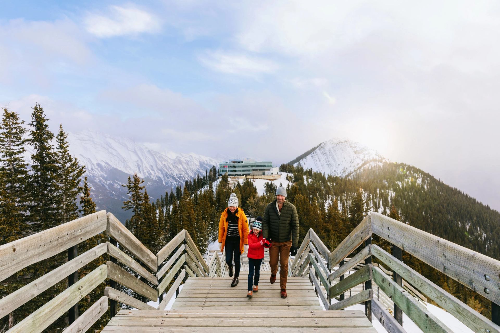 A family walking the boardwalk at Banff Gondola in Banff National Park. Mountains, trees and discovery centre in the background.