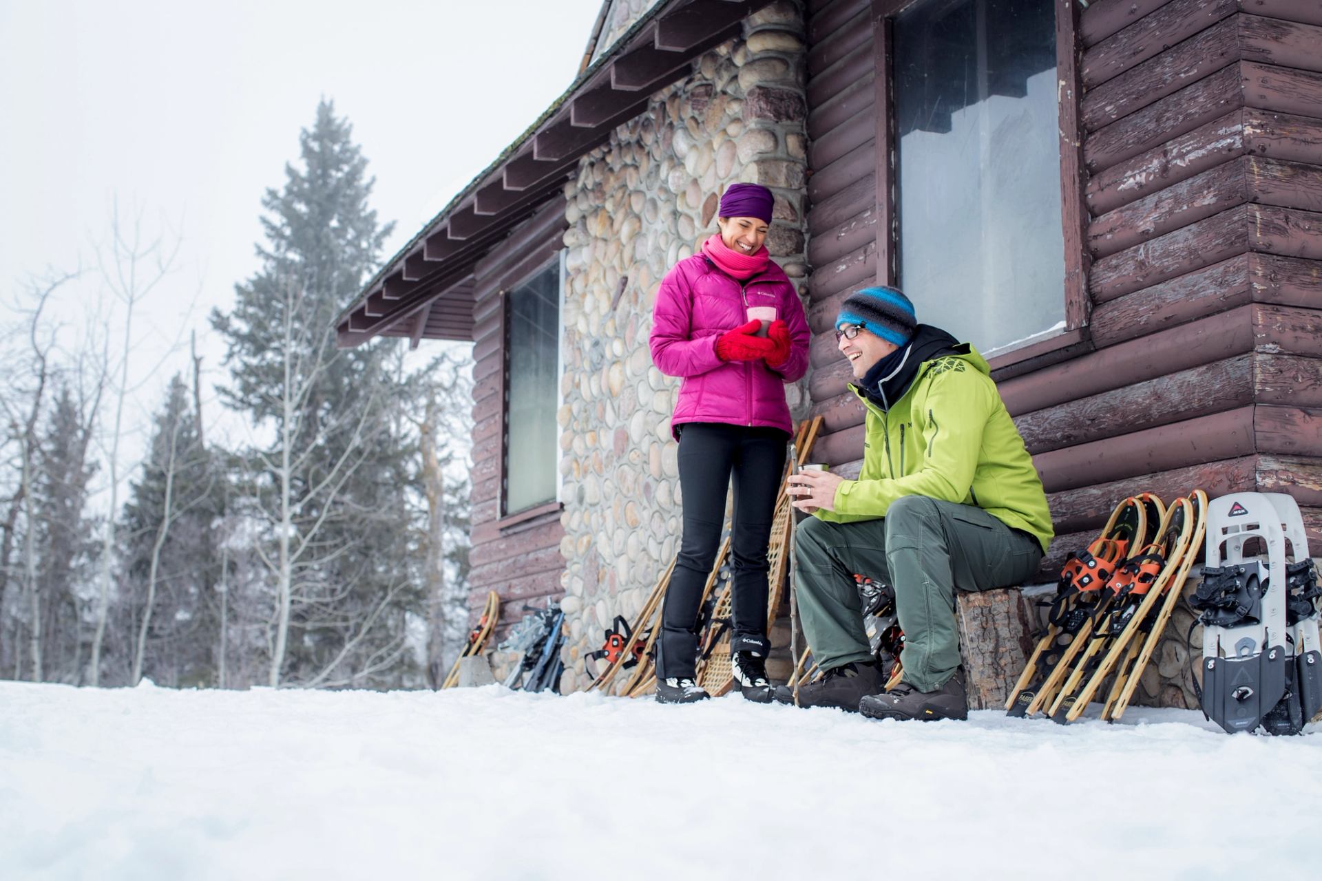 Couple with snowshoes having a hot drink outside a log cabin in Elk Island National Park