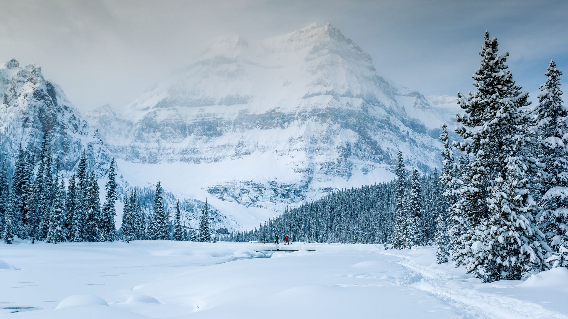 Two friends snowshoe across a bridge on their way into Shadow Lake in winter.
