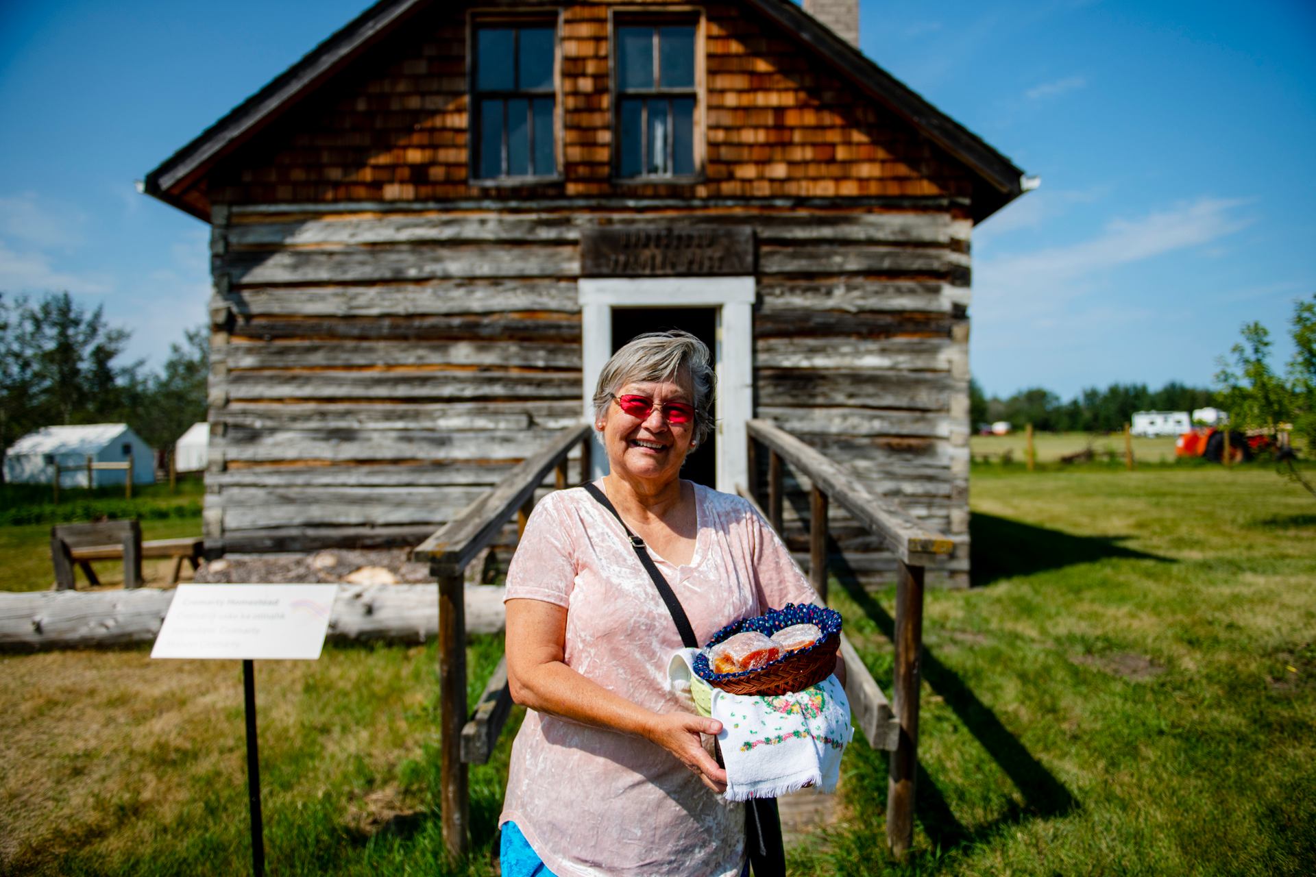 Indigenous elder woman holding basket of bannock bread in front of historic building at Metis Crossing.