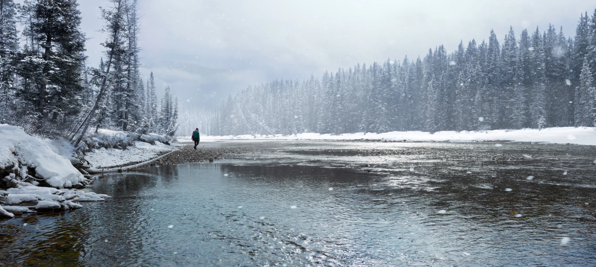 A woman walking along a glacier-fed river in Banff National Park while snow falls lightly.