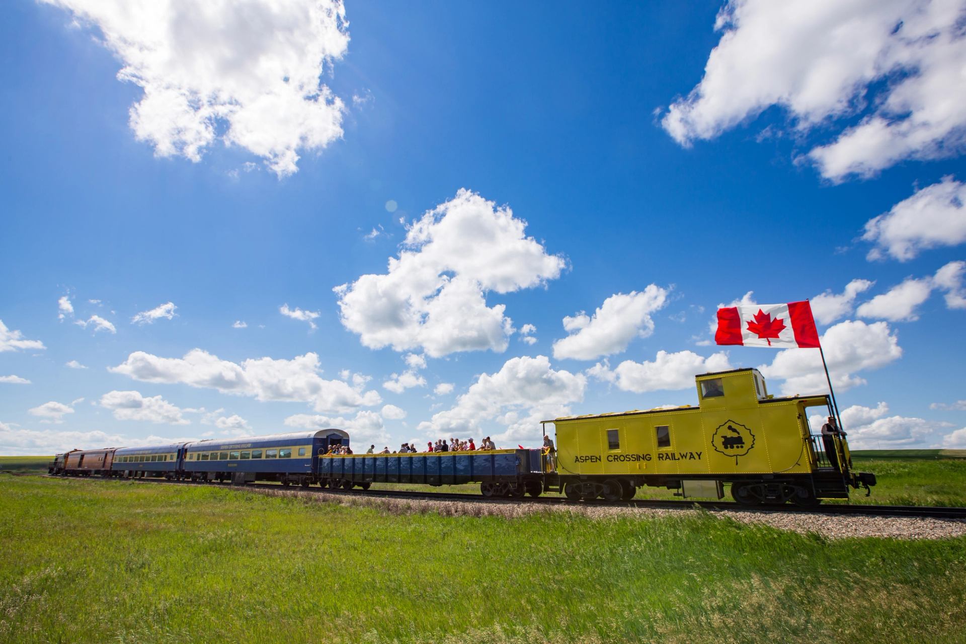 A Canadian flag waves on a yellow train car on the Aspen Crossing Train in the green prairies under a blue sky near Mossleigh.