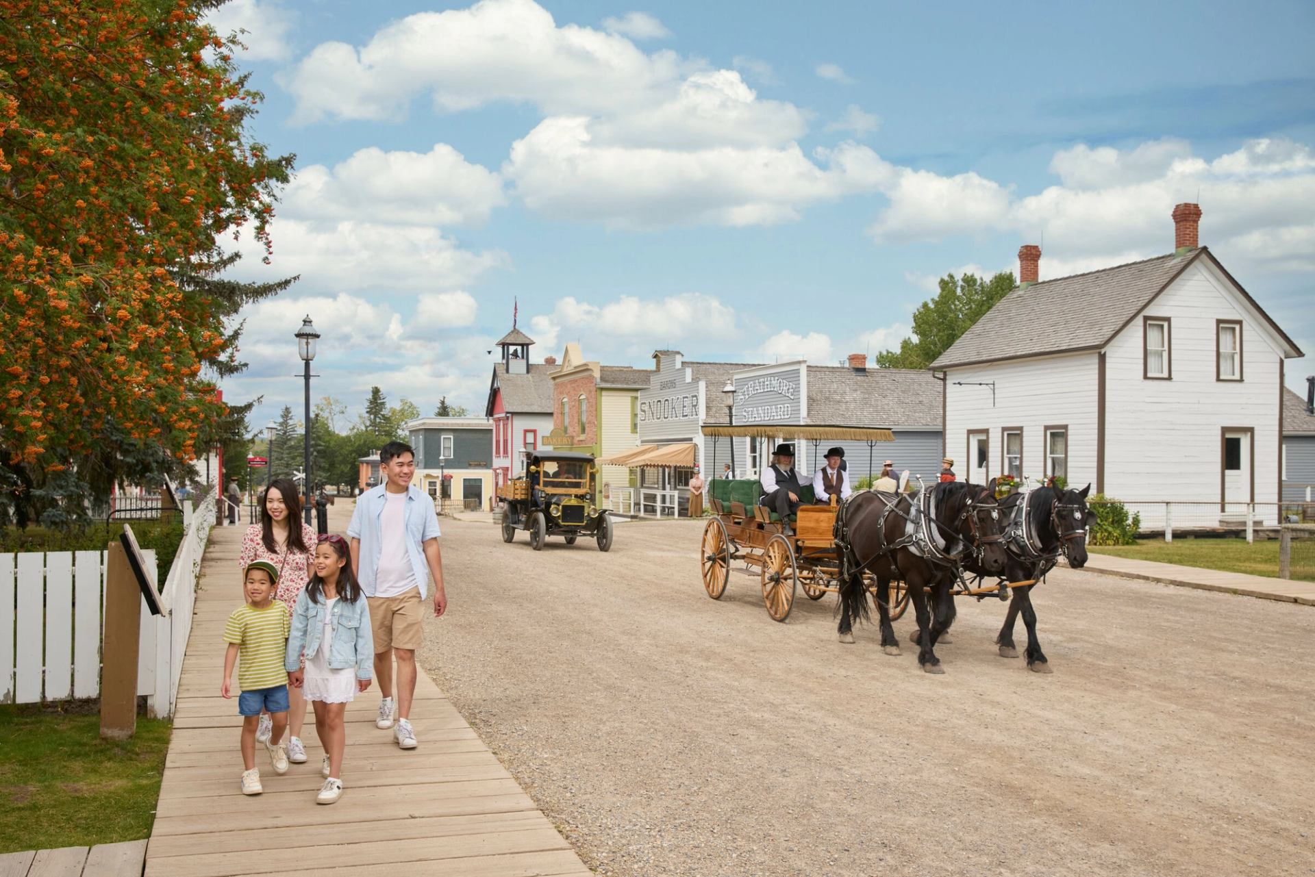 Family walks down a street in Heritage Park with a horse drawn carriage on the road.