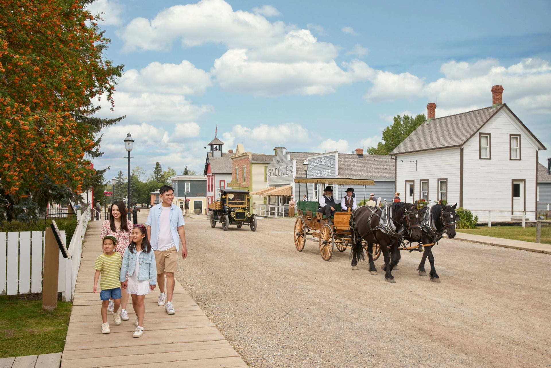 Family walks down a street in Heritage Park with a horse drawn carriage on the road.