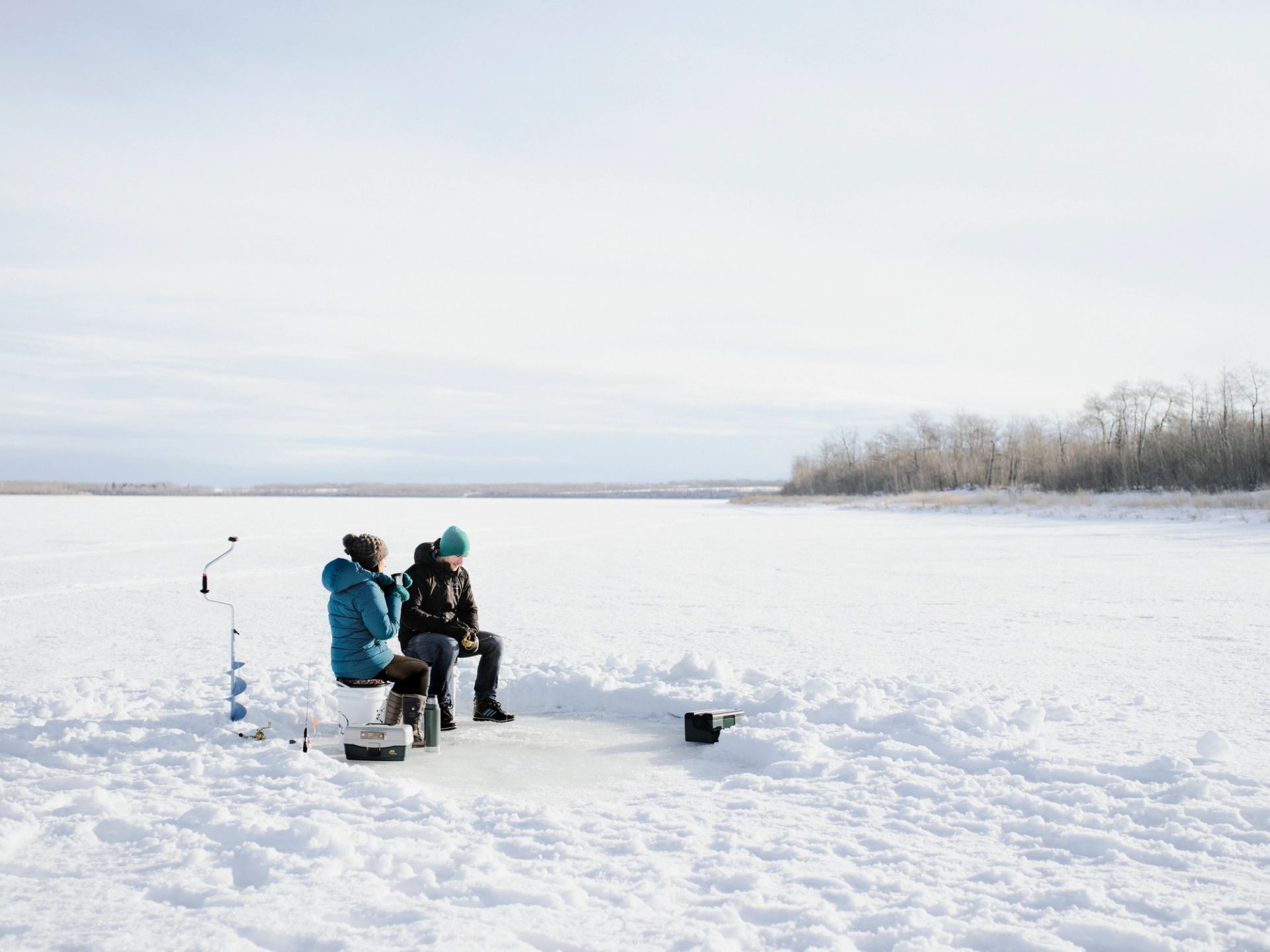 A couple ice fishing and drinking hot beverages while sitting on a a frozen Bear Lake near Grande Prairie.