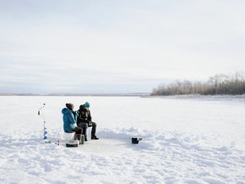 A couple ice fishing and drinking hot beverages while sitting on a a frozen Bear Lake near Grande Prairie.