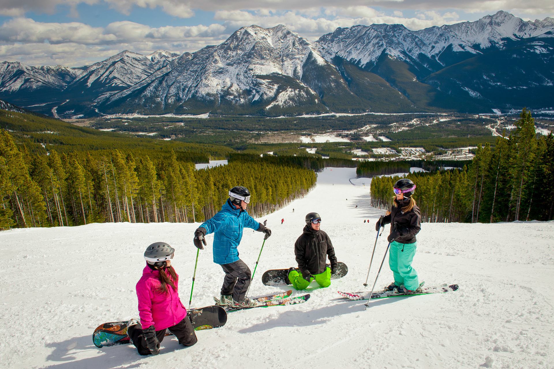 Four skiers and snowboarders take a break on the hill at a ski resort with mountains in the background.