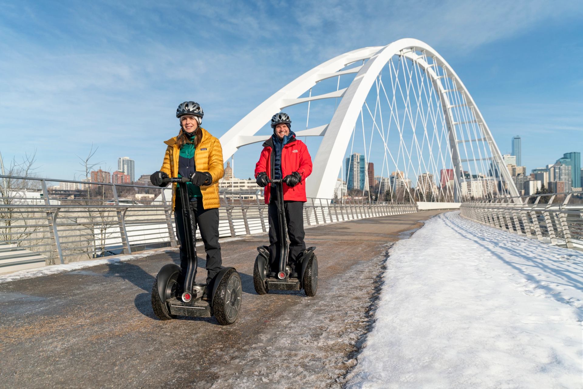 Two people go over a bridge on a Segway in winter with a ciy in the background.