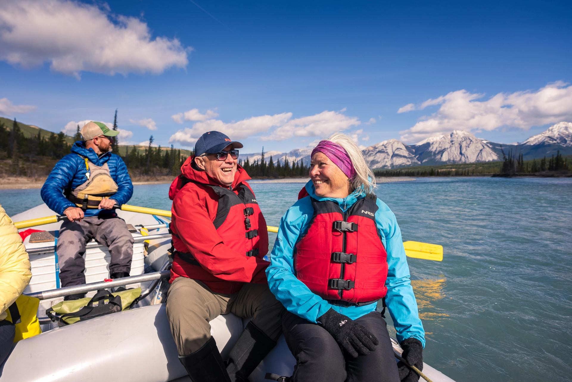 A group in a raft floating down a calm river with forested banks and mountains in the distance.